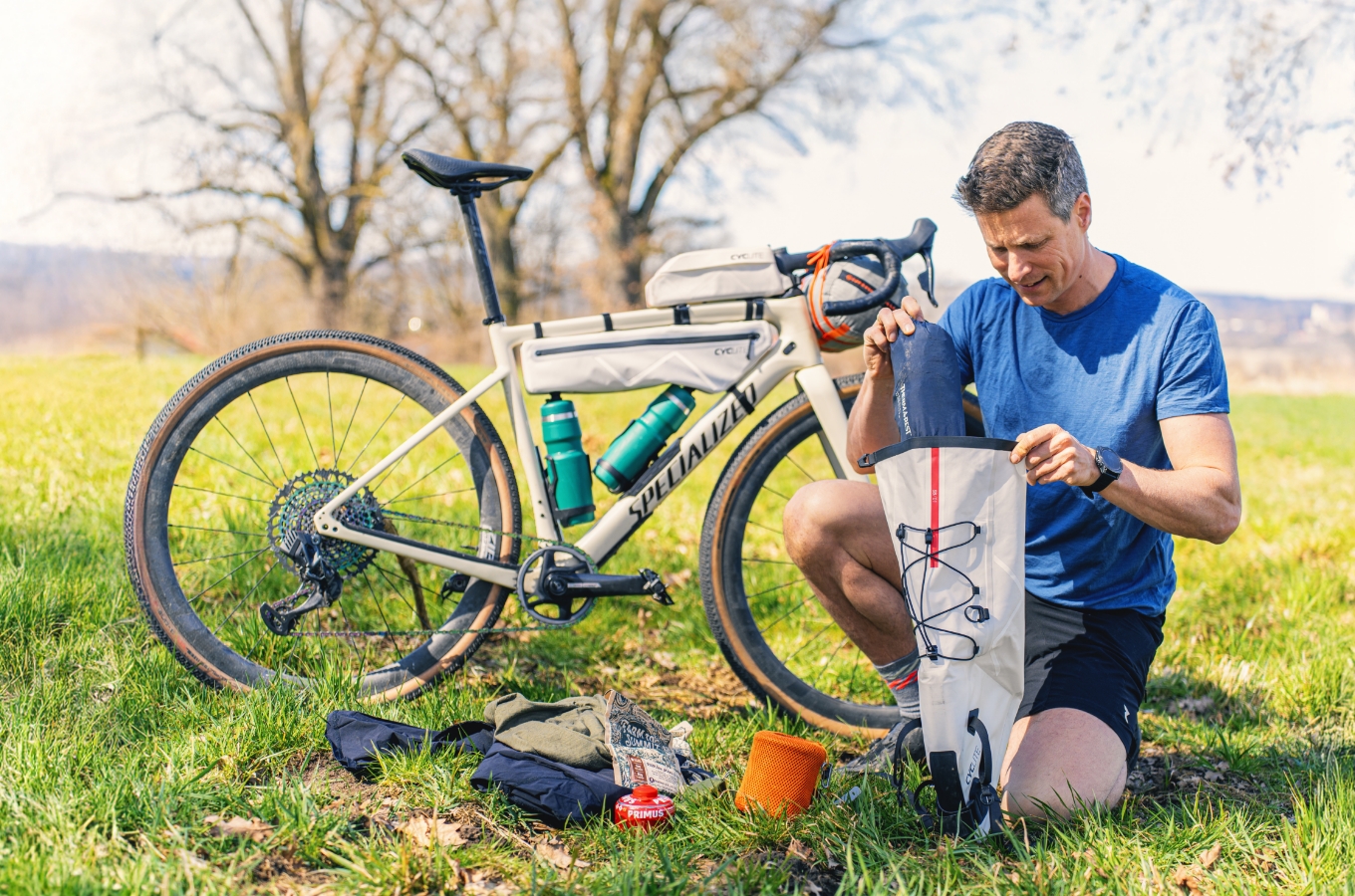 Bikepacker packt Ausrüstung in Tasche, Gravelbike und Equipment auf einer Wiese ausgelegt.